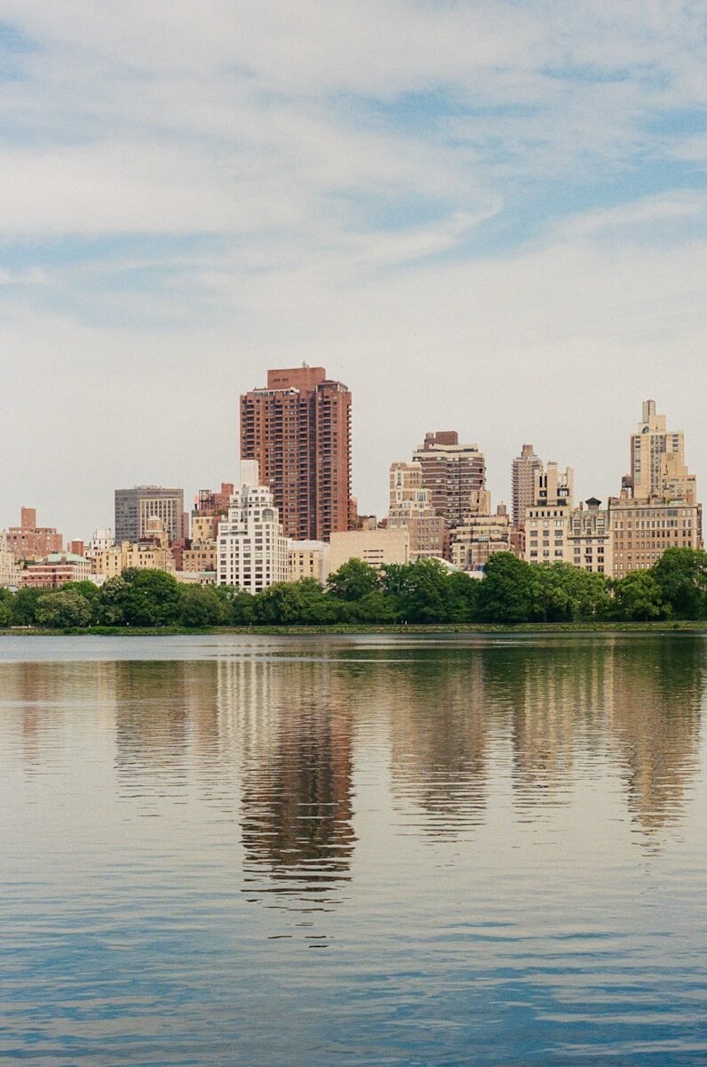 Skyscrapers and trees reflected in tranquil water under cloudy sky.